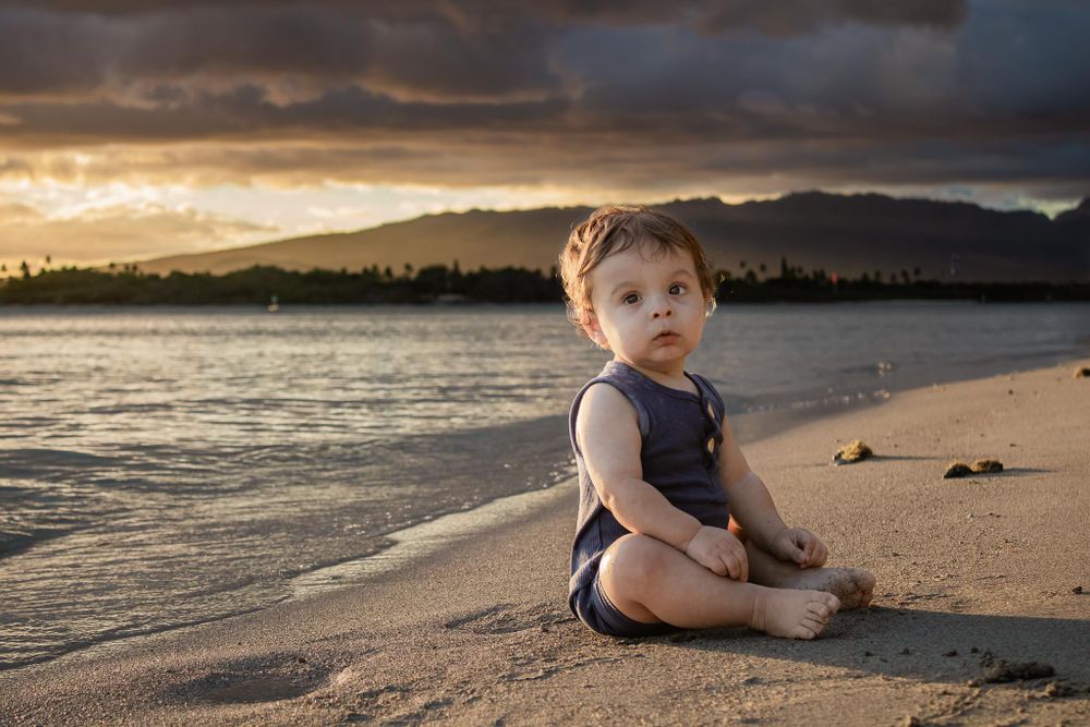Toddler in navy romper sitting on beach during dramatic sunset.