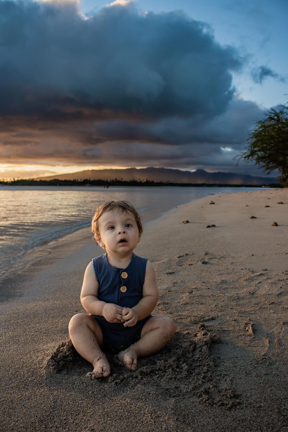 A small child in a navy blue romper sits on a sandy beach at sunset with dramatic clouds and mountains in the background.
