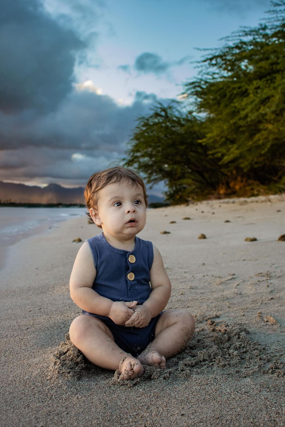 A toddler in a blue outfit sits on a tropical beach at dusk with moody clouds overhead.