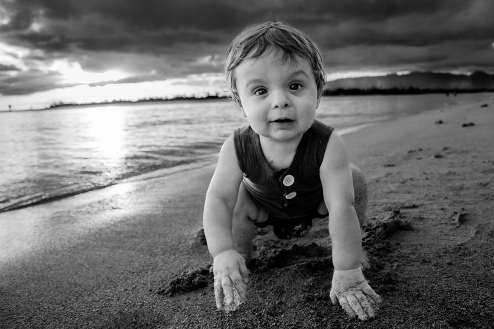 A black and white photo of a baby crawling on a beach at sunset with dramatic stormy skies.
