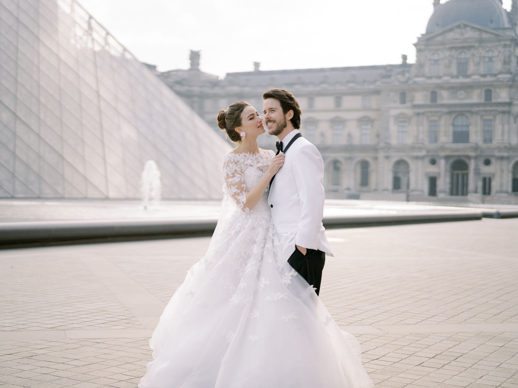 A romantic wedding portrait in front of the Louvre pyramid on a misty day.