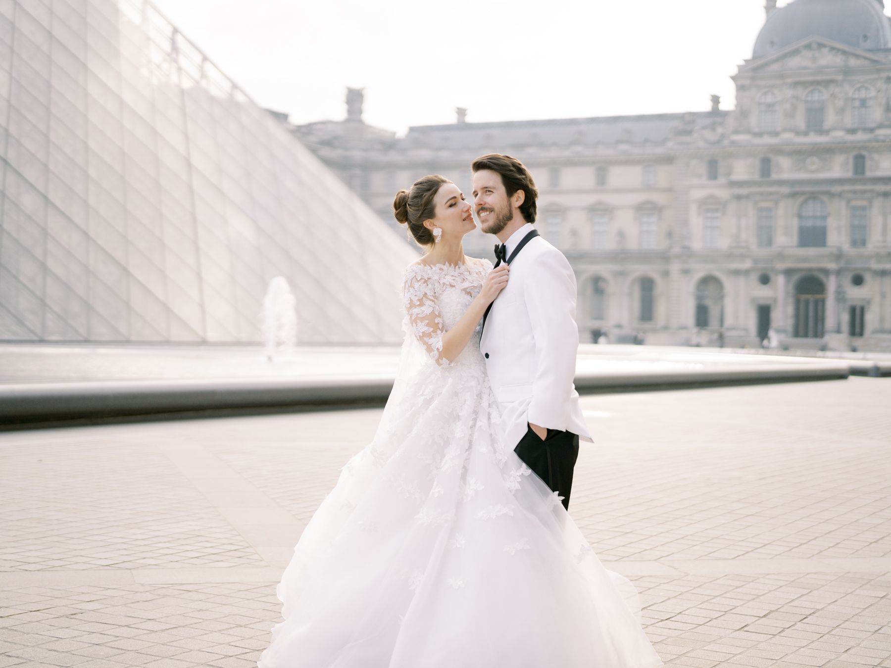 A romantic wedding photo of a couple embracing by the Louvre Pyramid in Paris.