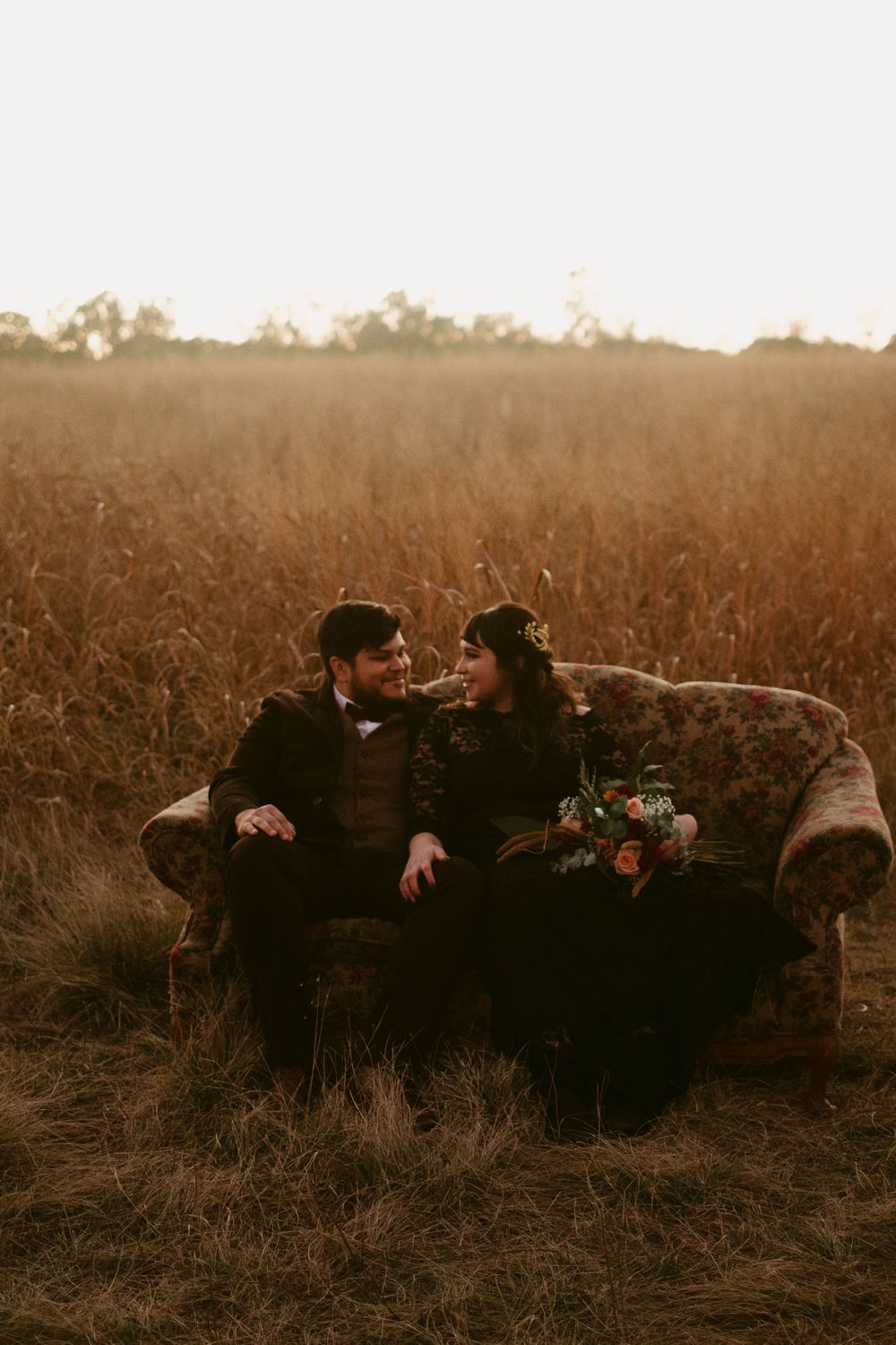 Two people sit together on an antique sofa placed in a rustic field during golden hour.