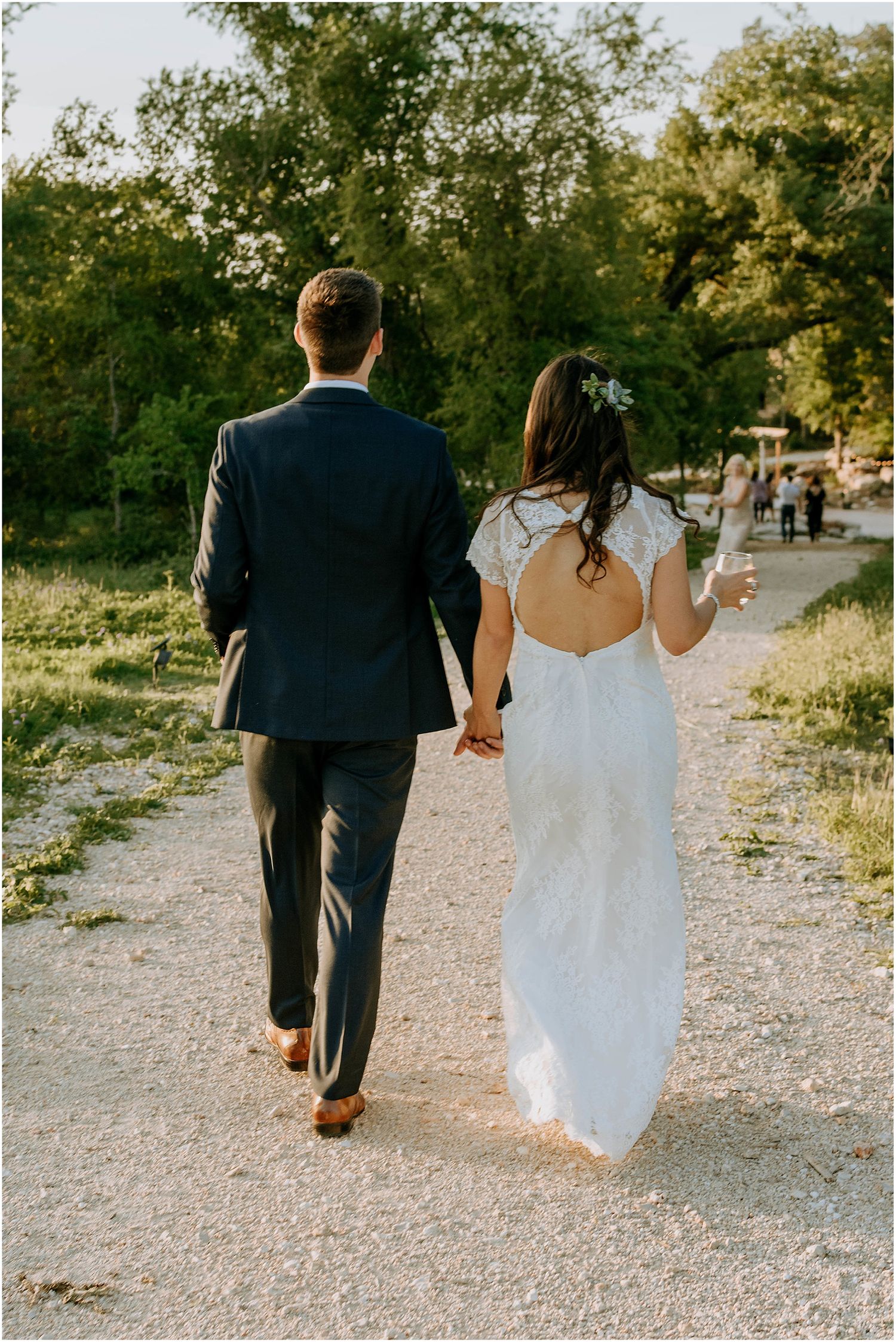 Couple walking hand in hand down a gravel path in wedding attire at sunset.