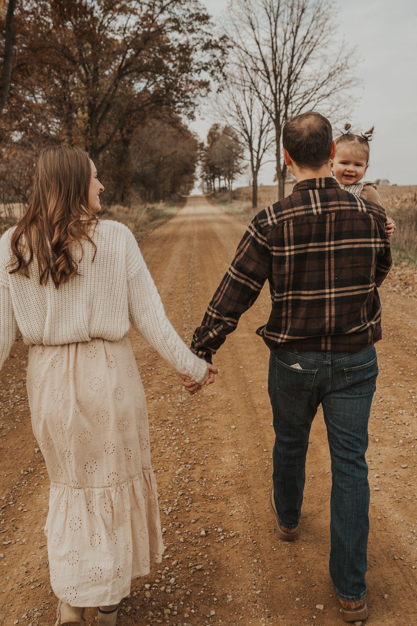 A couple and child walk hand in hand down a rustic dirt path surrounded by autumn trees near Jackson Michigan.