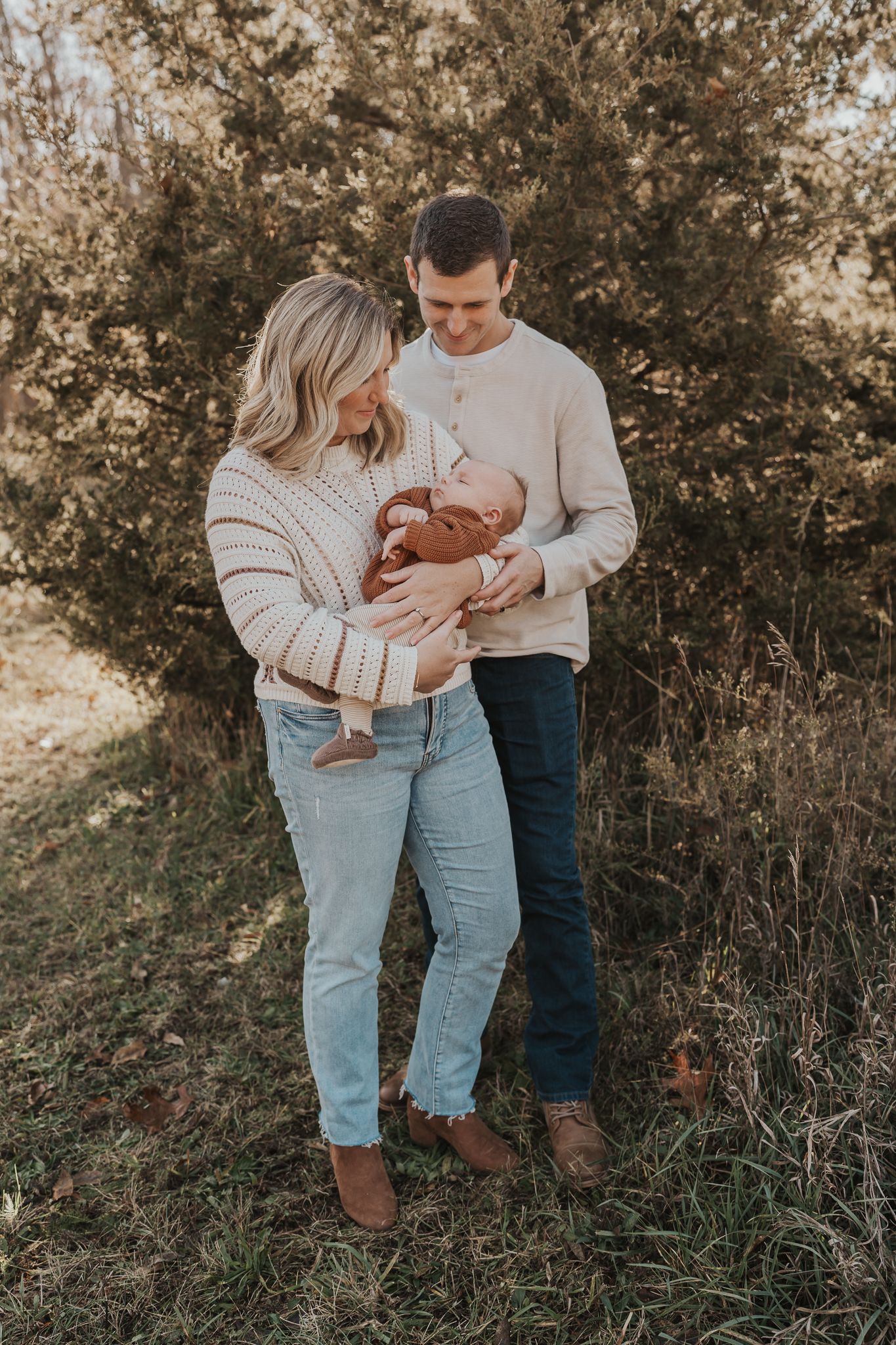A couple in casual denim and sweaters embraces while holding their baby outdoors.