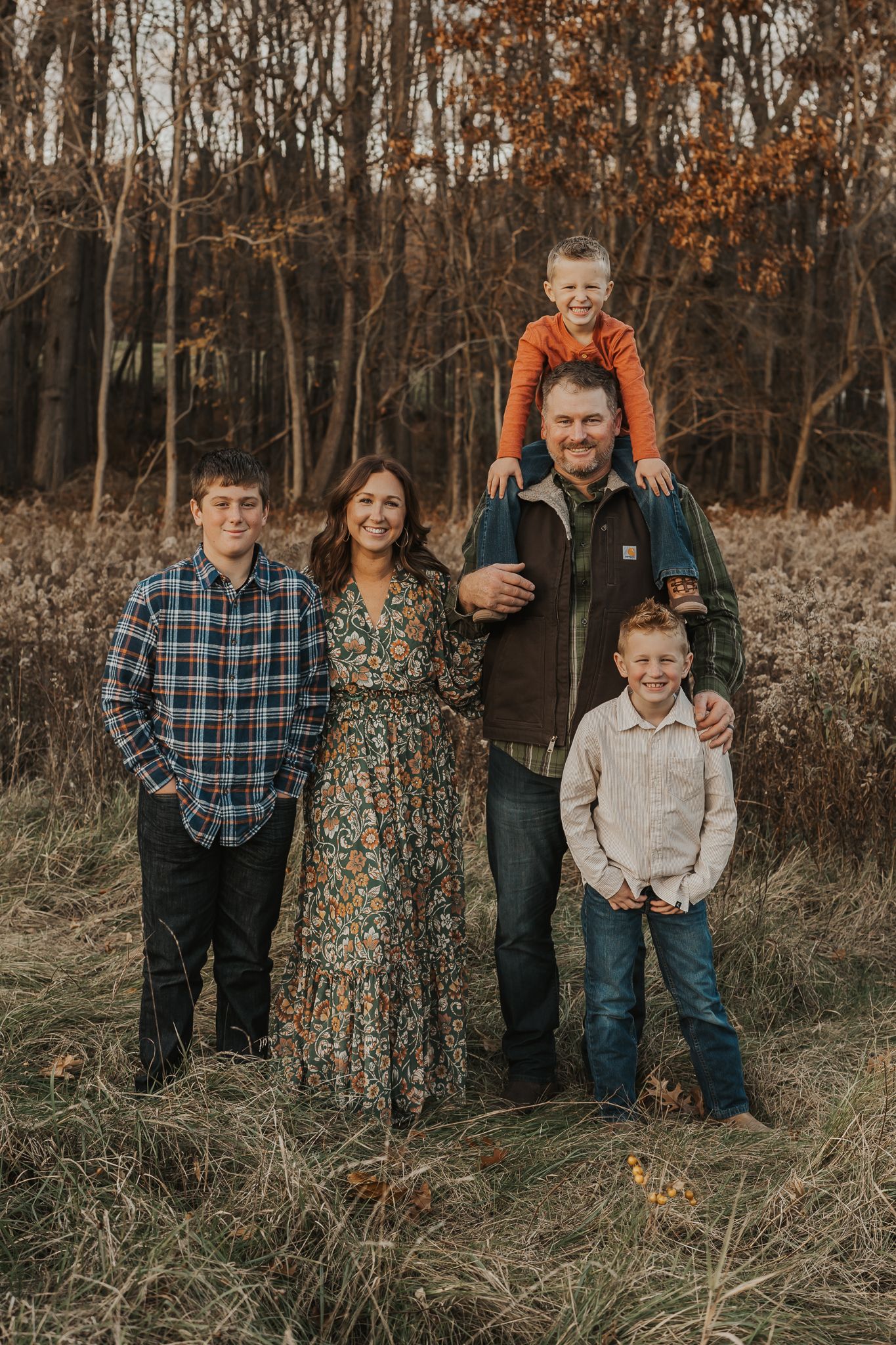 A family wearing coordinated fall outfits poses together in a rustic outdoor setting with autumn foliage.