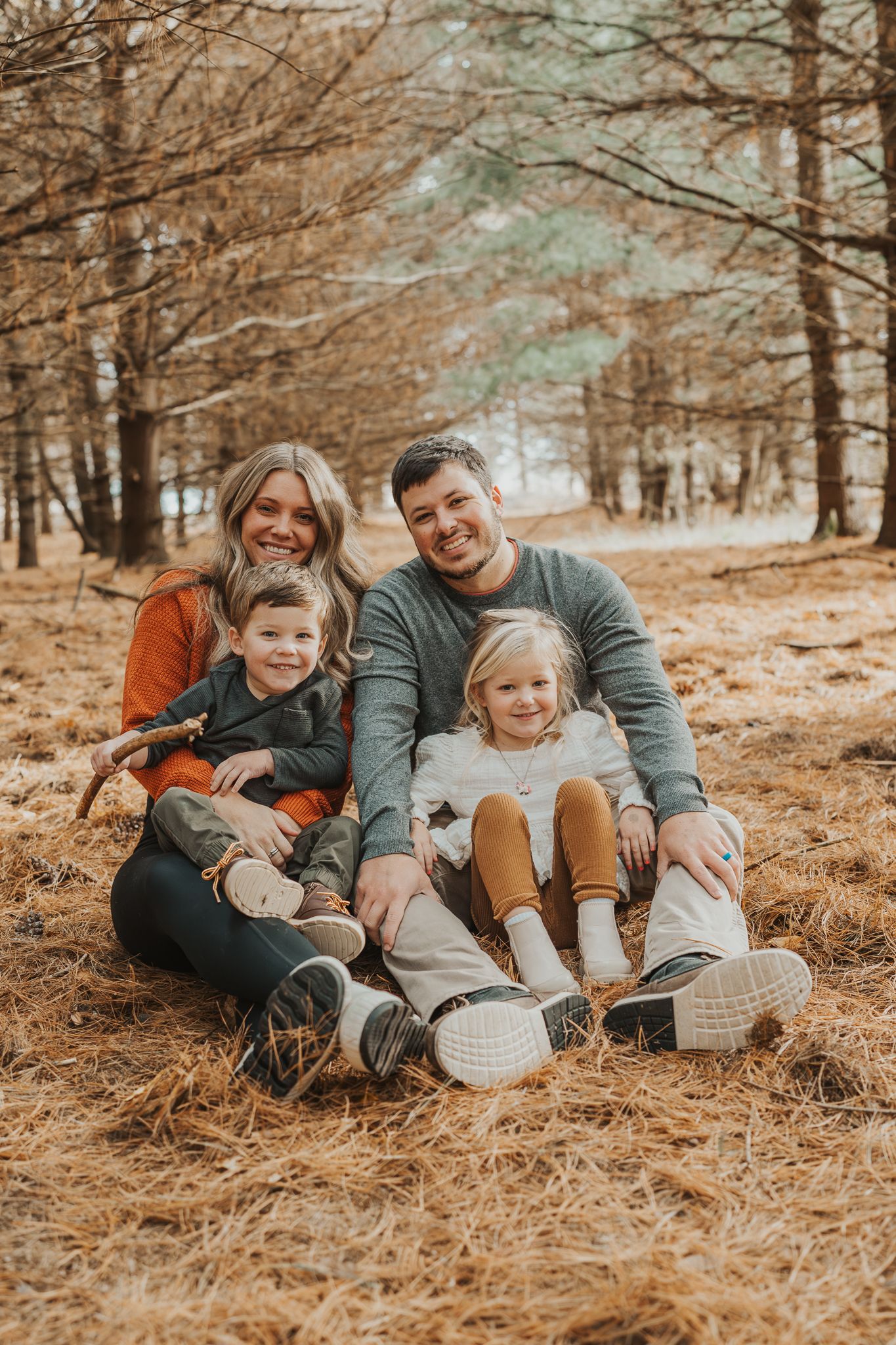 A happy family sits together on fallen pine needles in an autumn forest setting.
