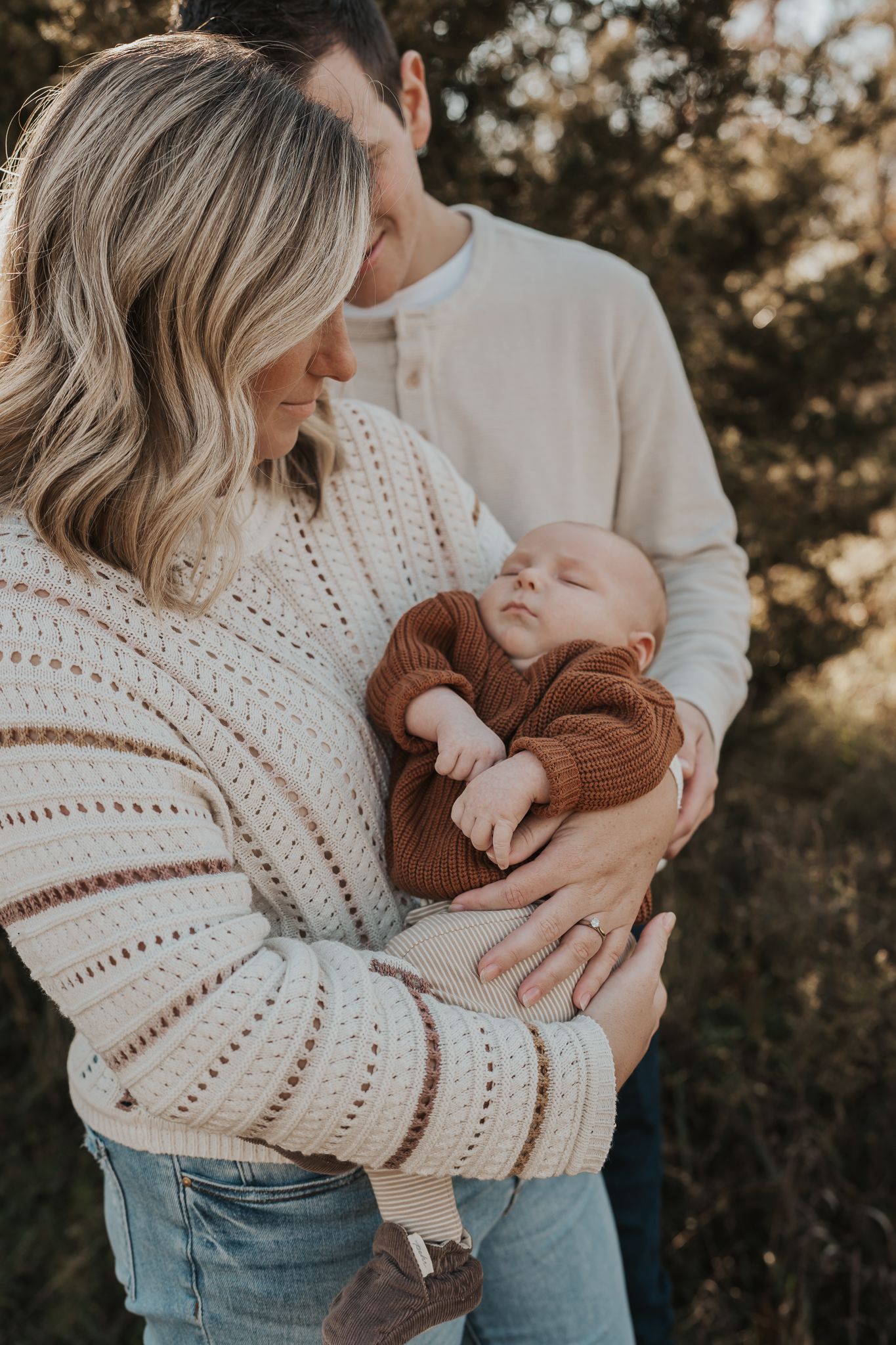 Parents in neutral sweaters cradle their baby wearing a brown knit outfit during a photo session taken by a Michigan photographer.