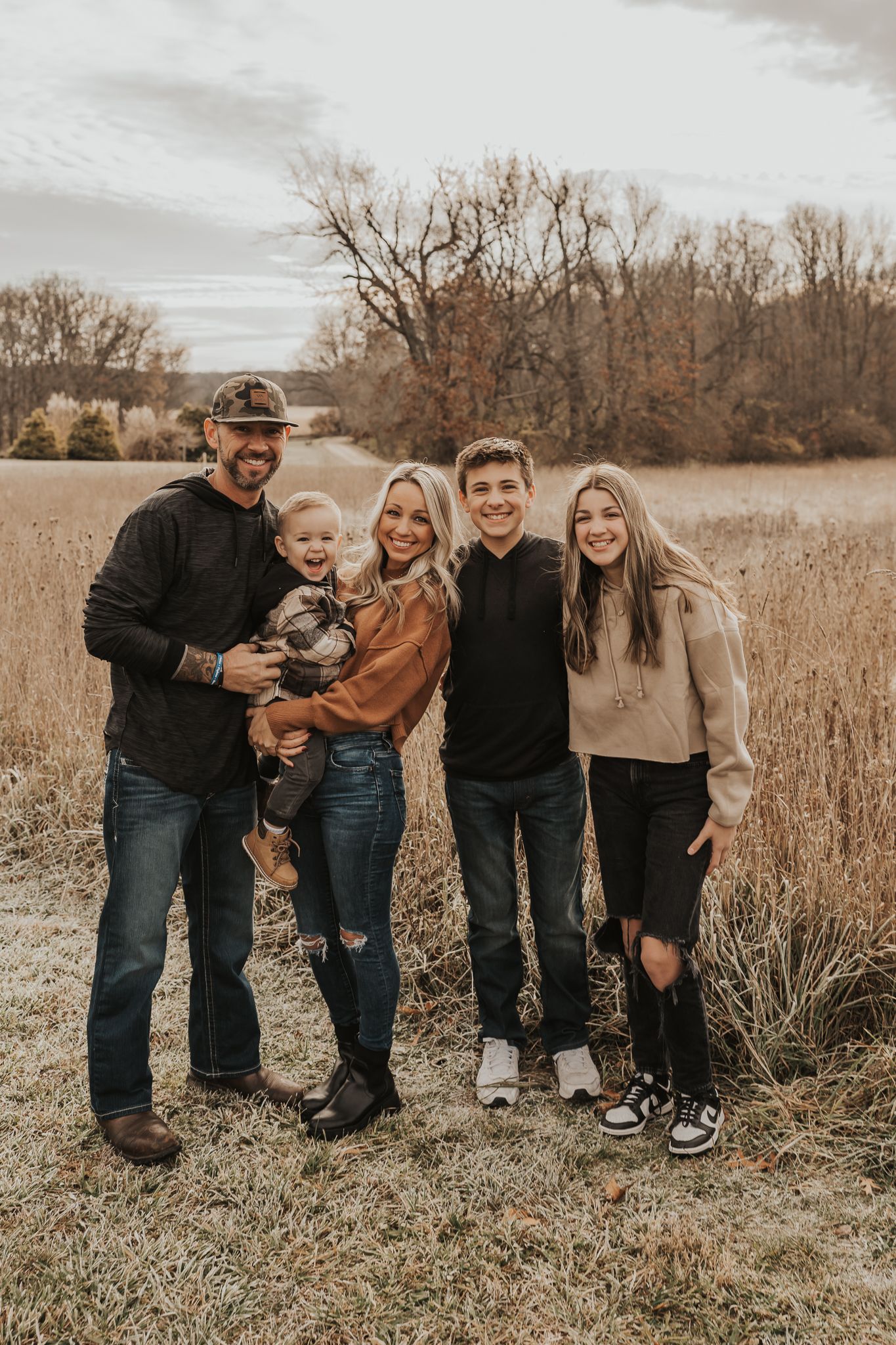 Family wearing coordinated casual outfits poses together in a rustic autumn field setting.