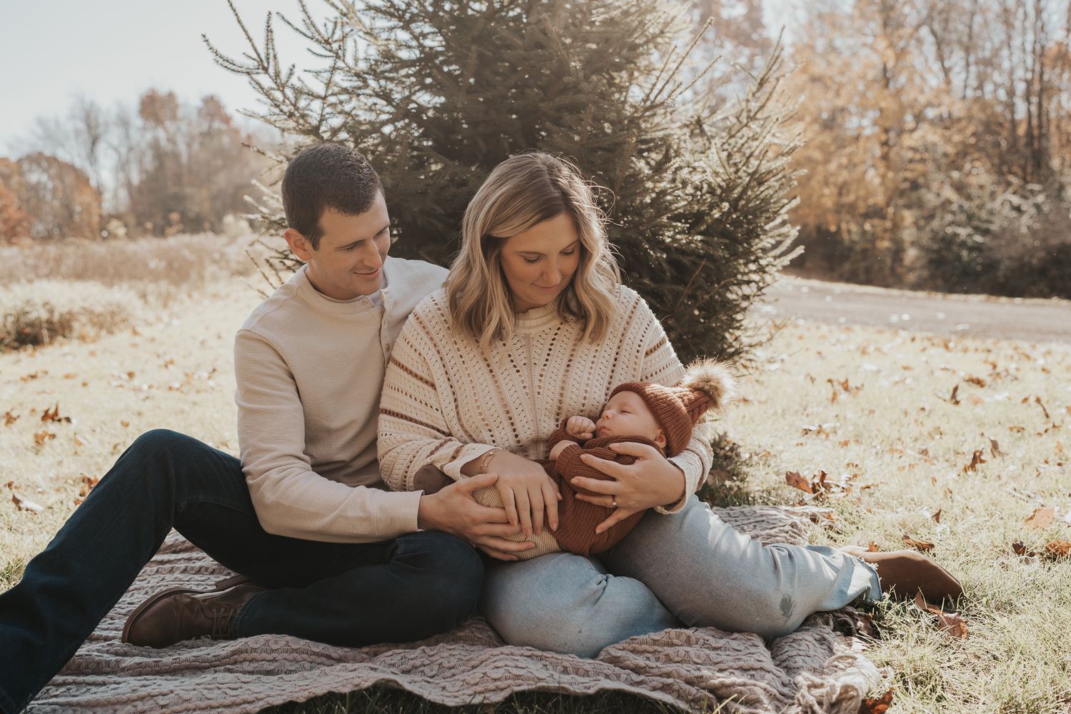 A couple sits together on a blanket outdoors while holding their infant.