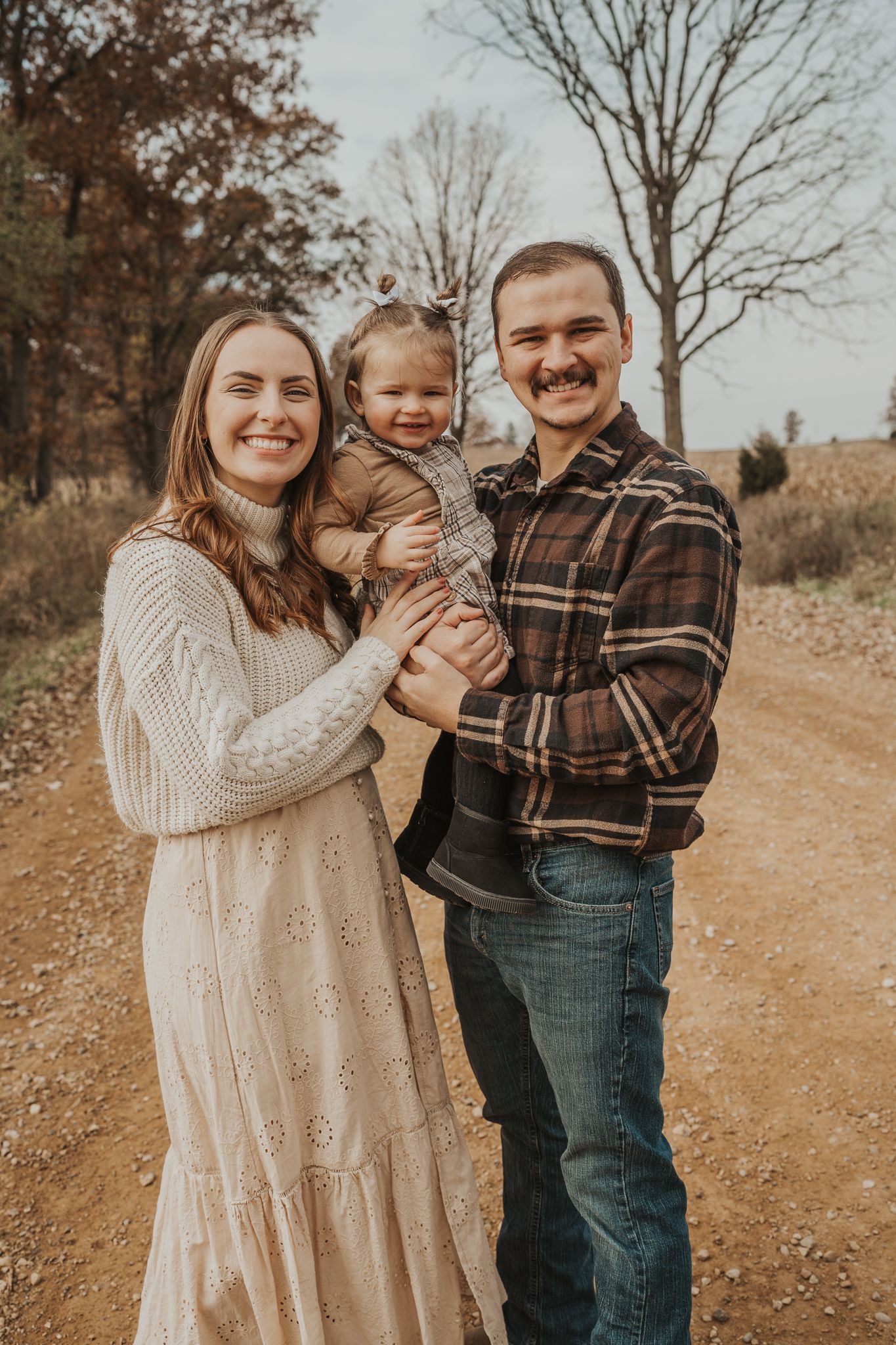 A family poses together on a dirt path surrounded by autumn trees in warm earth tones.