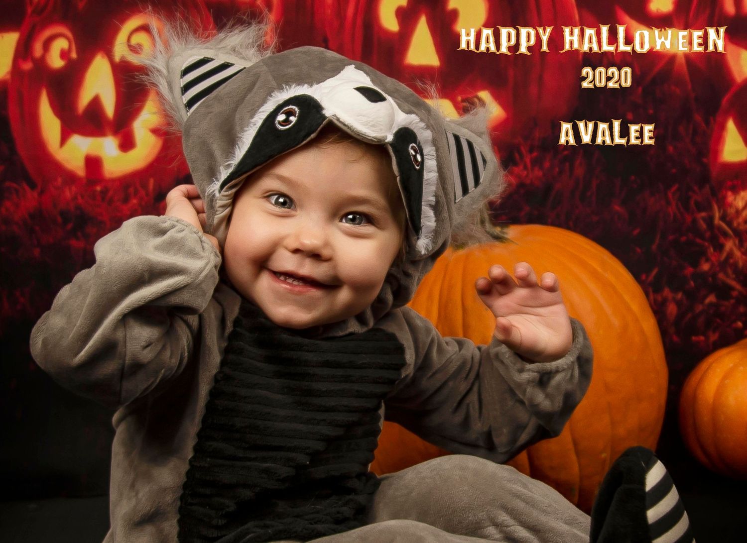 A small child in a grey animal costume poses with pumpkins against a backdrop of glowing jack o'lanterns.