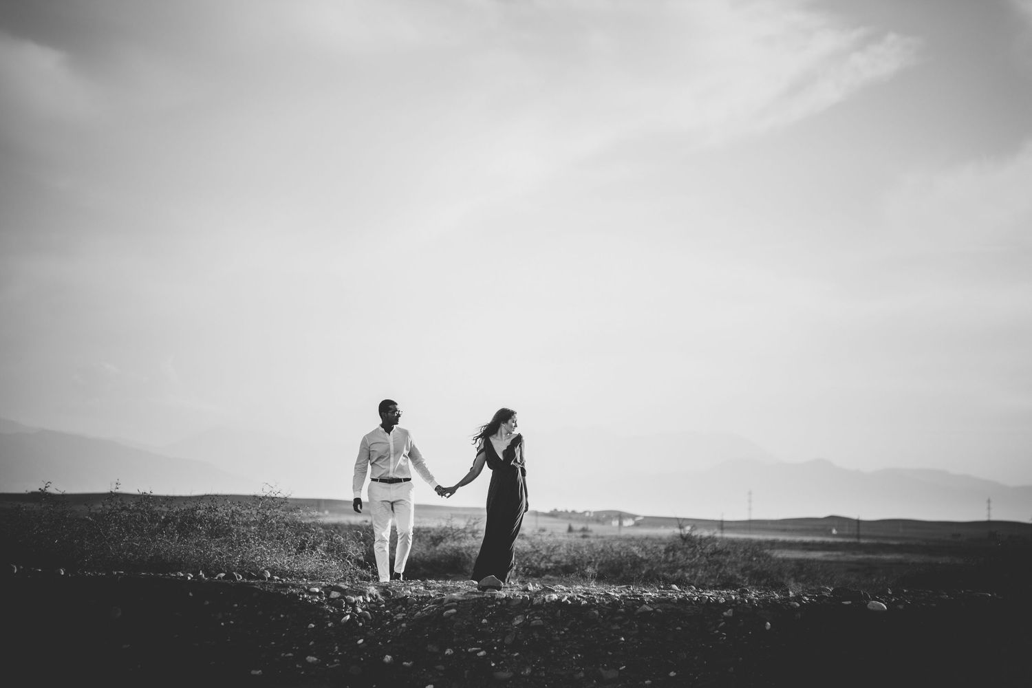 A couple stands together on a scenic hilltop overlooking a vast landscape in black and white.