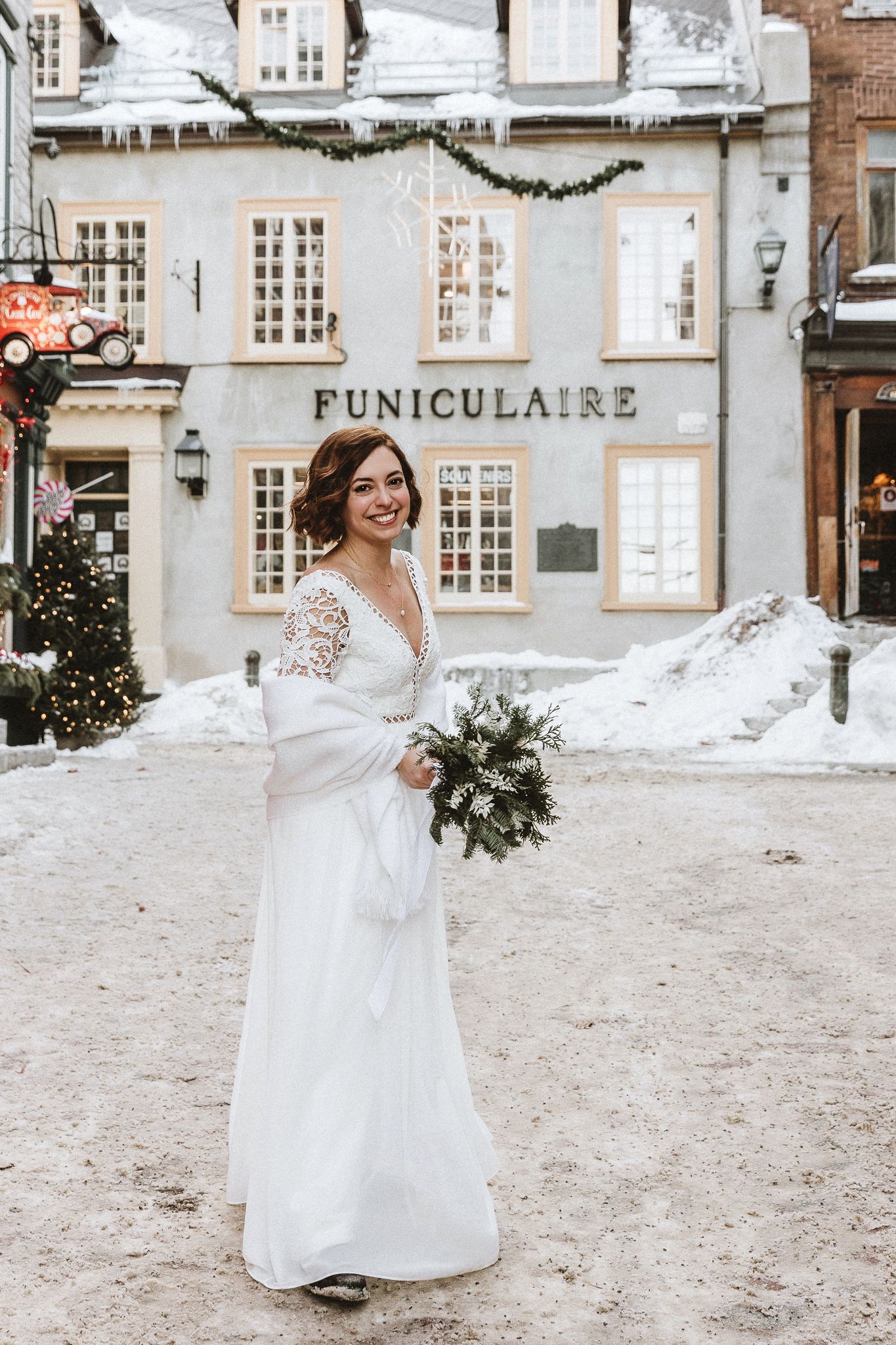 A winter bride in a lace wedding dress poses in front of the Funiculaire building in snow.