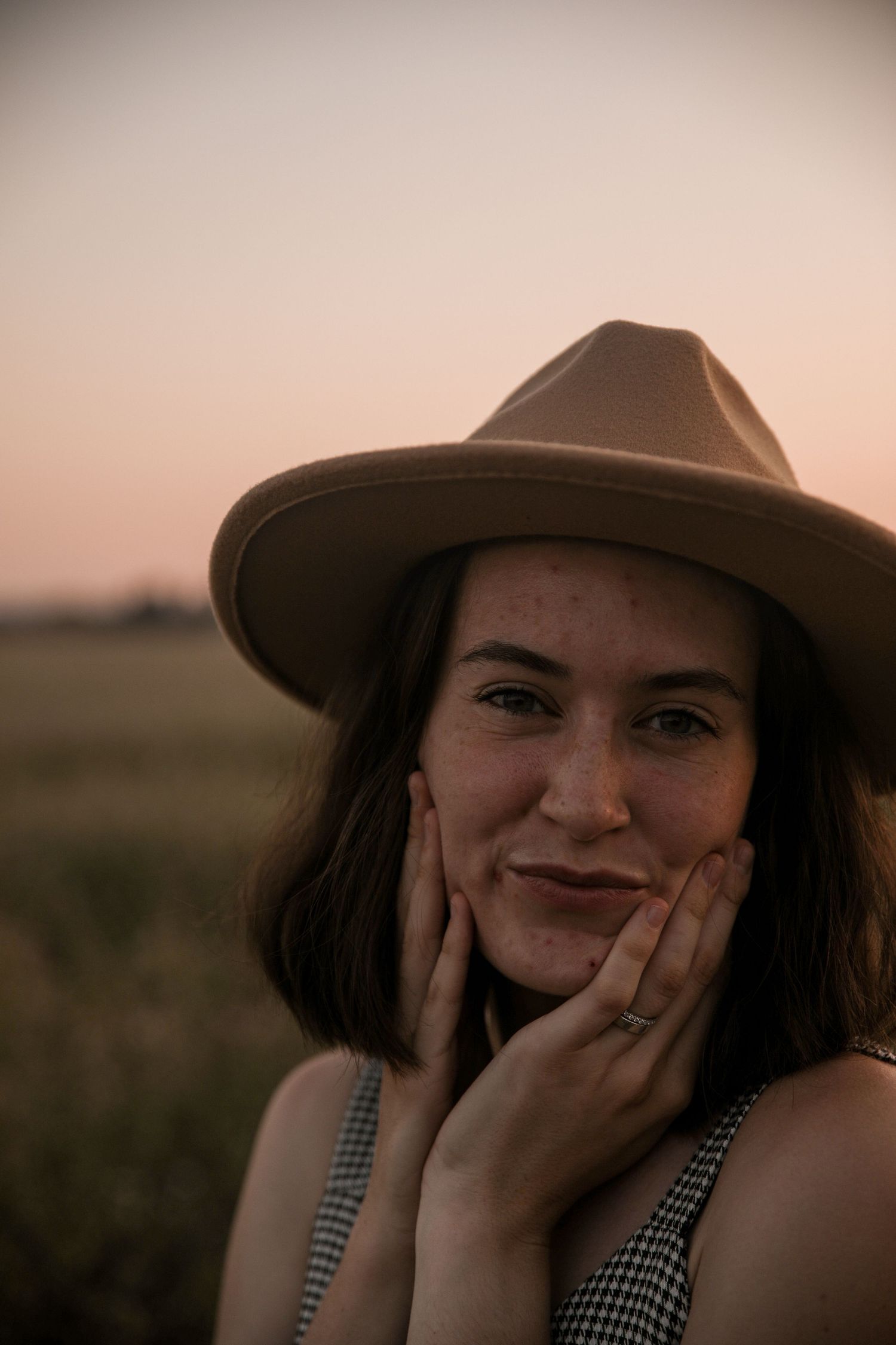 Portrait in warm sunset light with brown fedora and gingham dress in rural setting.