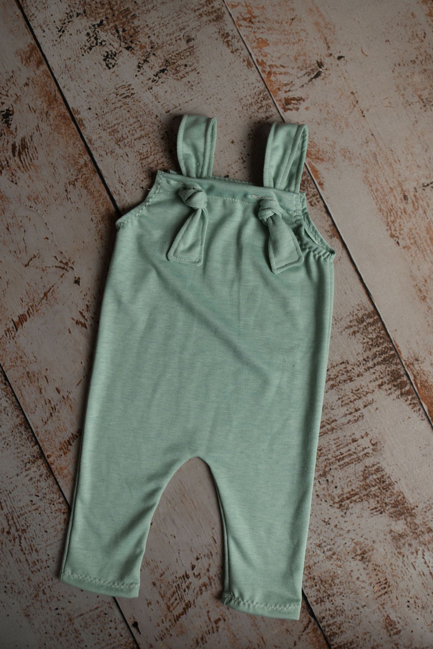 Sage green baby overalls with tie details laid flat on distressed wooden surface.