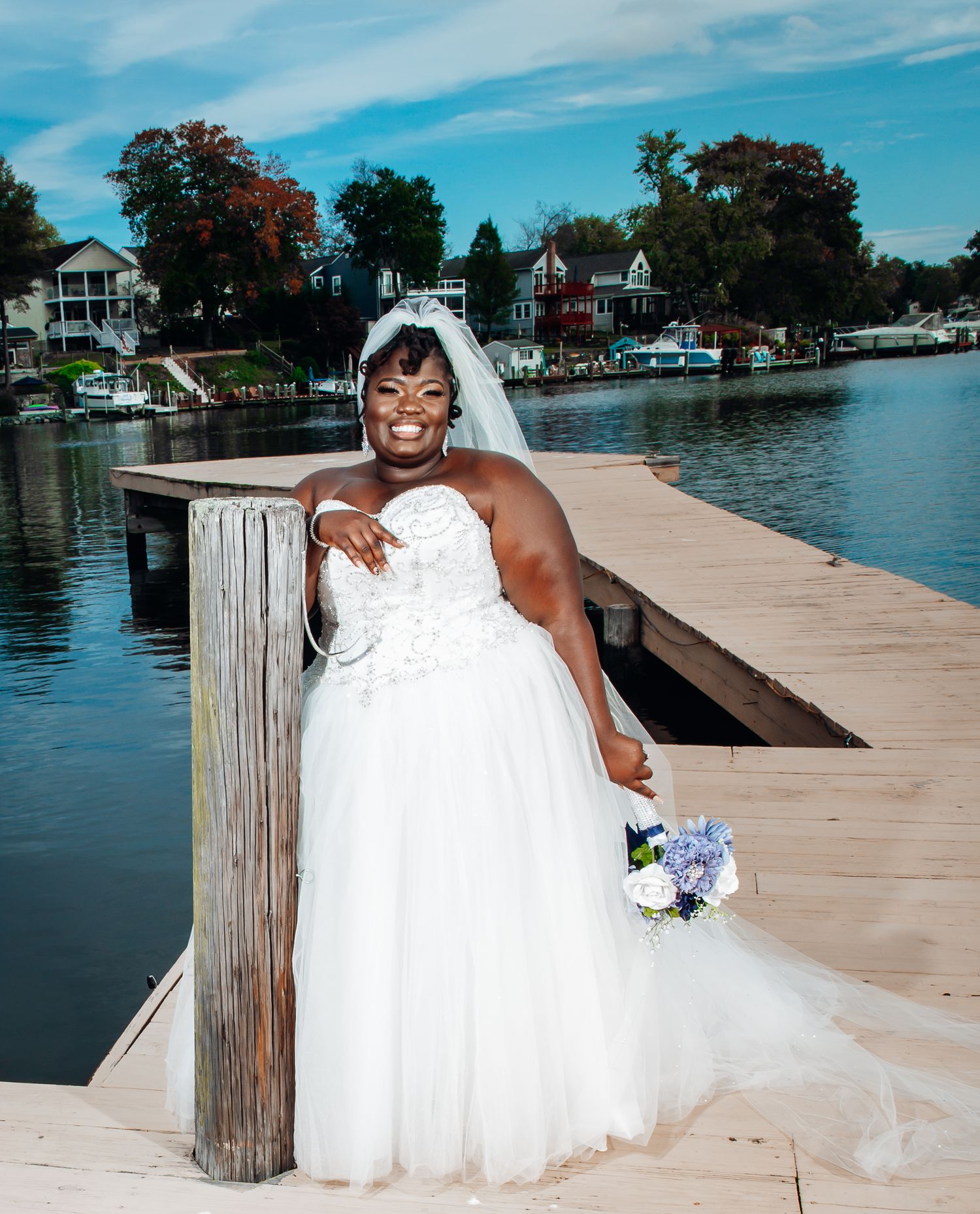 A bride in a white strapless ball gown and veil poses on a wooden dock beside a lake holding flowers.