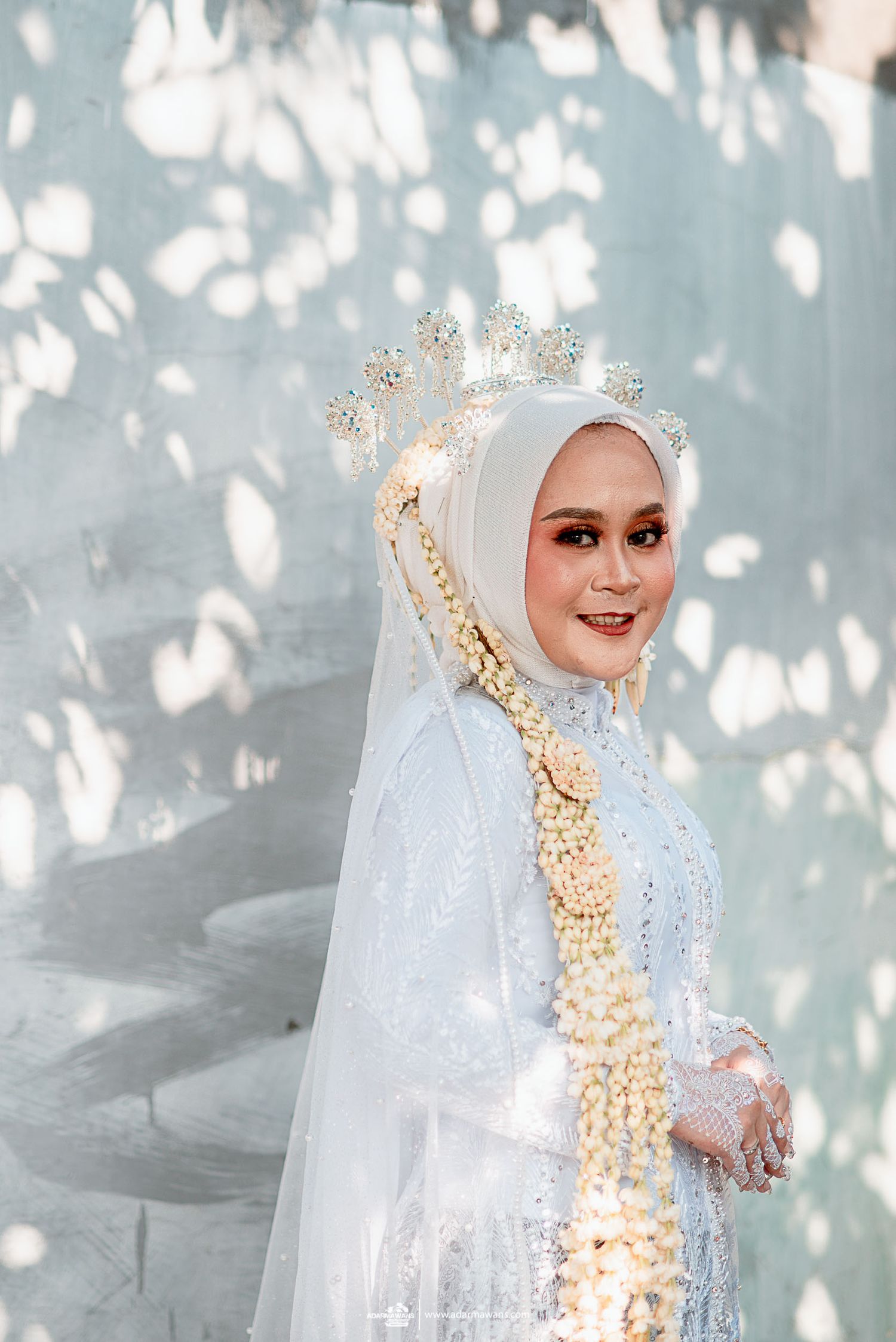 Elegant bride in white hijab and ornate crown poses against dappled light background during traditional ceremony.