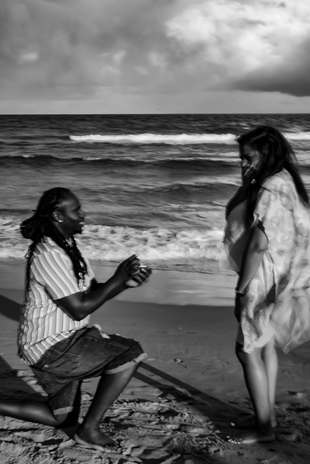 Black and white beach proposal photo of man kneeling before woman at the ocean shore.