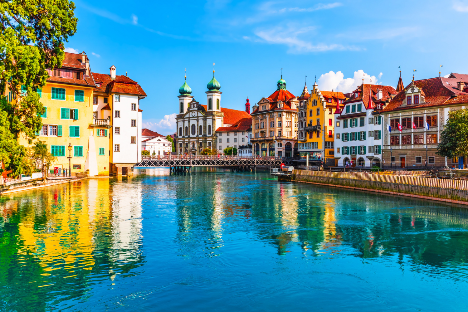 Colorful historic buildings along the Reuss River waterfront in Lucerne, Switzerland reflect in turquoise waters on sunny day.