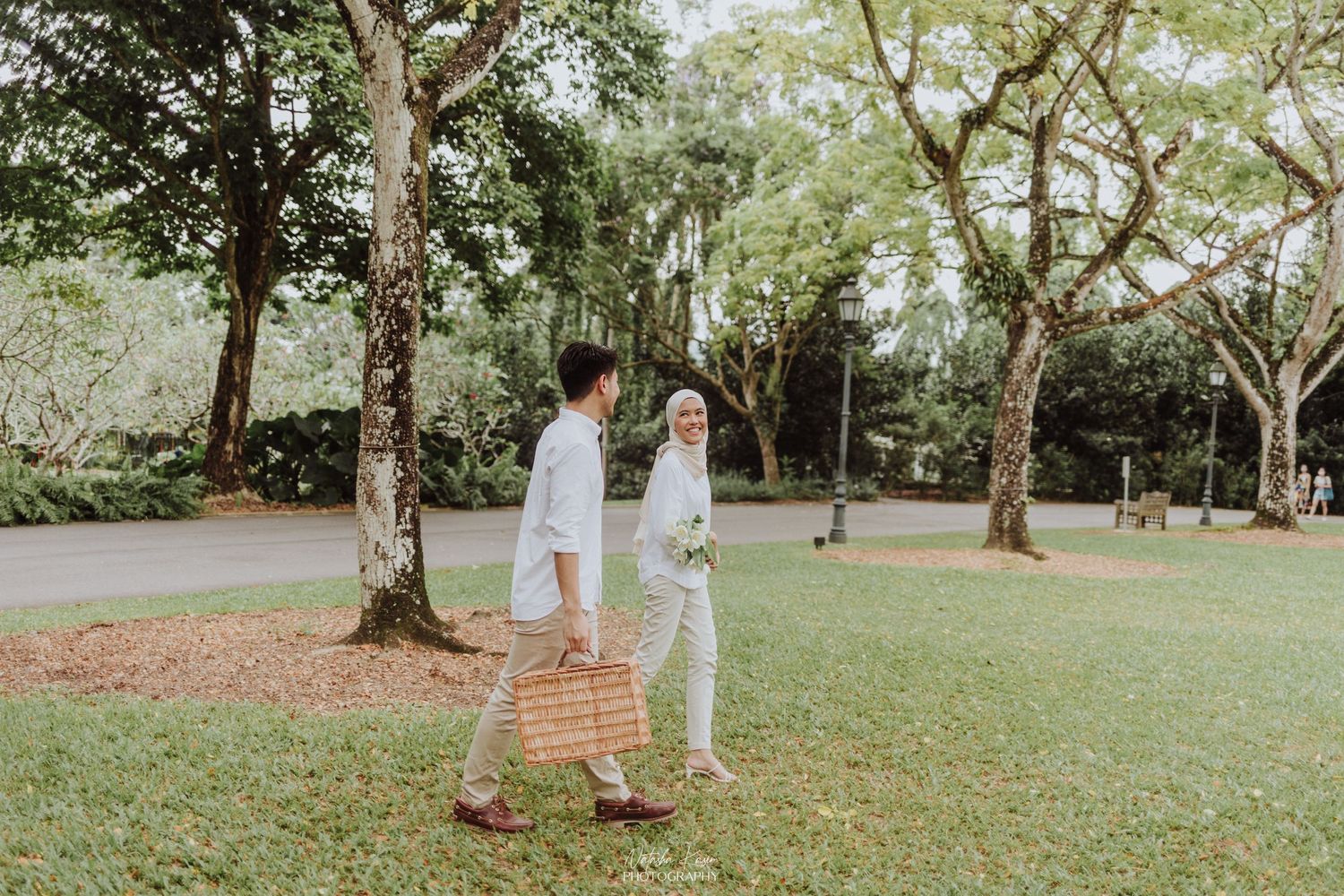 A couple takes a romantic stroll through a park with tall trees on a sunny day.