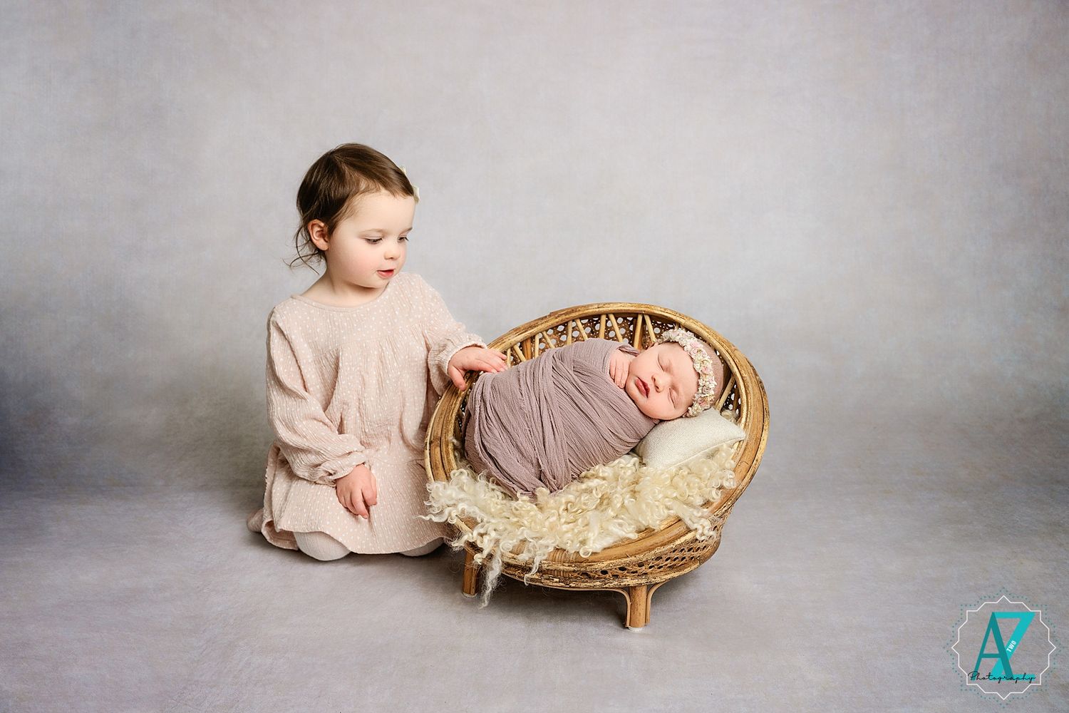 Child in beige dress looking at basket with baby on grey background.