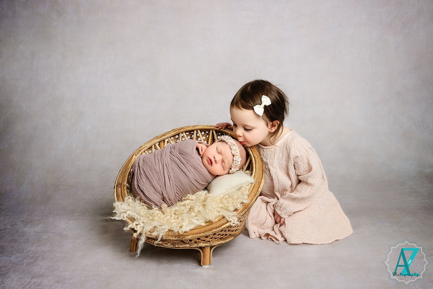 Two young children pose for a heartwarming portrait with one resting in a rustic basket filled with hay.