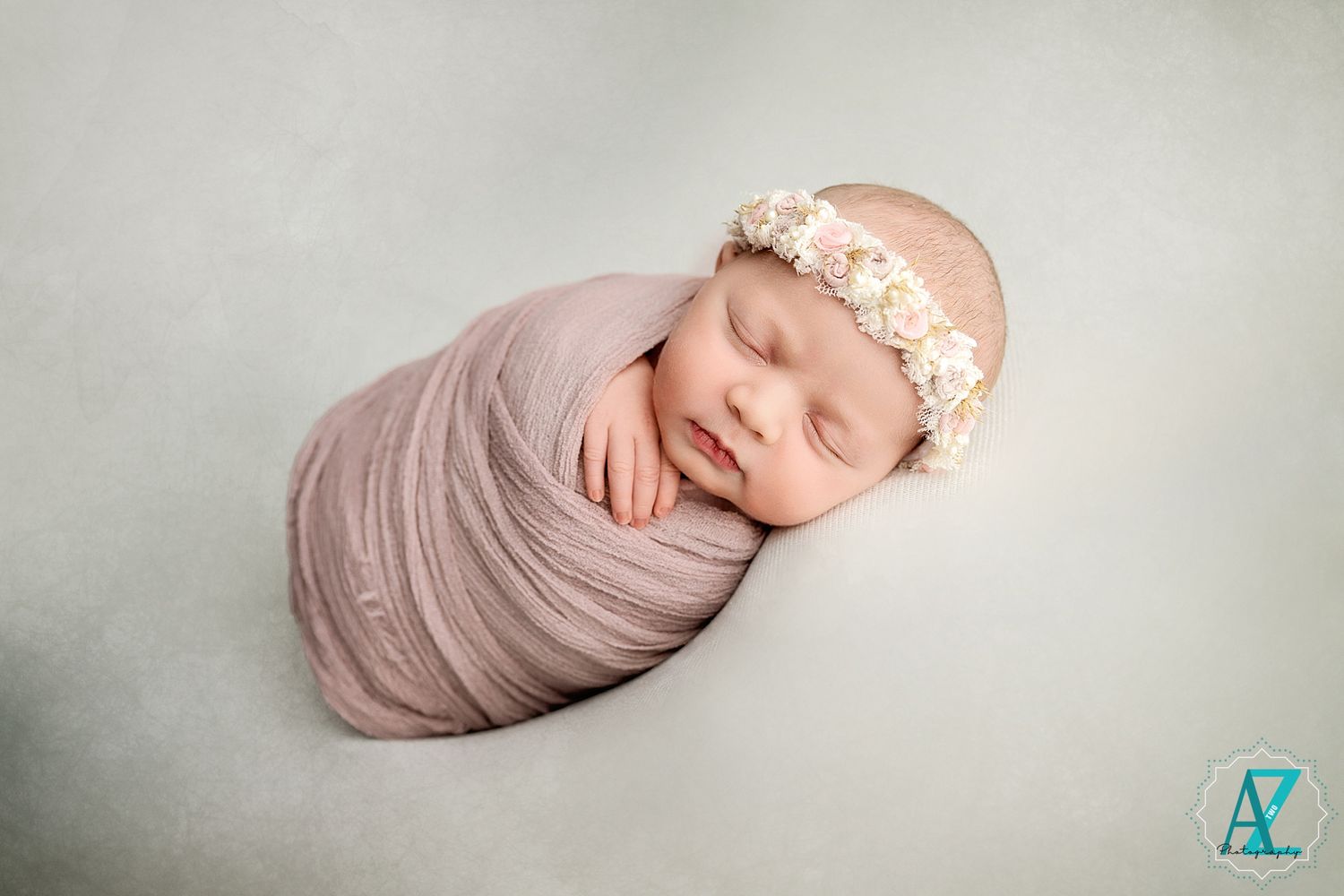 Newborn baby wrapped in pink swaddle wearing floral headband against neutral background.