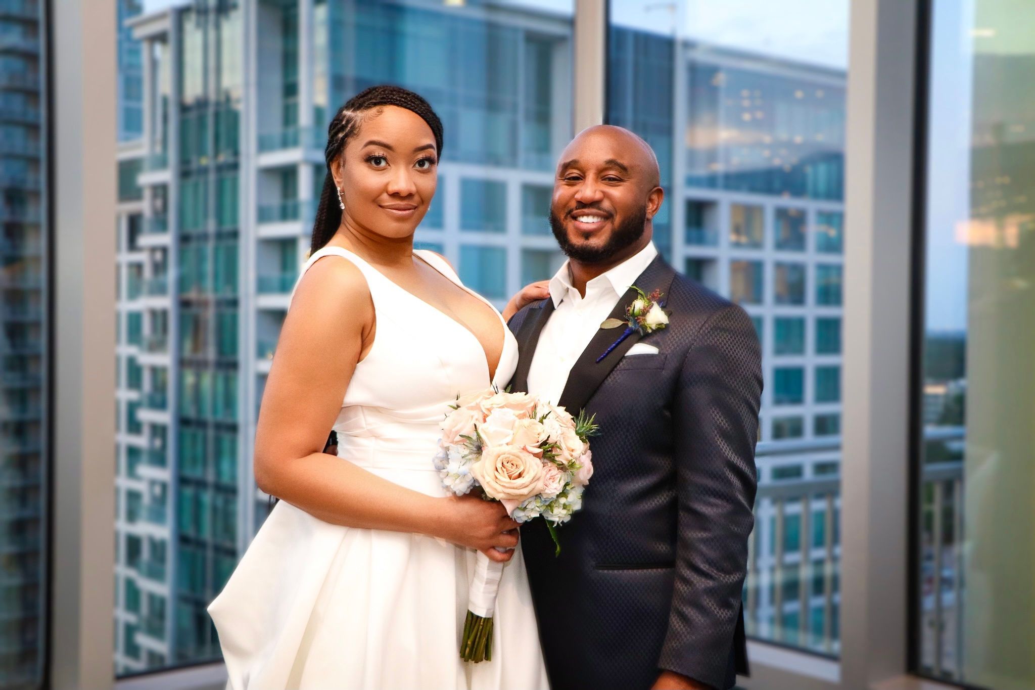 Happy couple in wedding attire pose with bouquet against modern glass building backdrop.