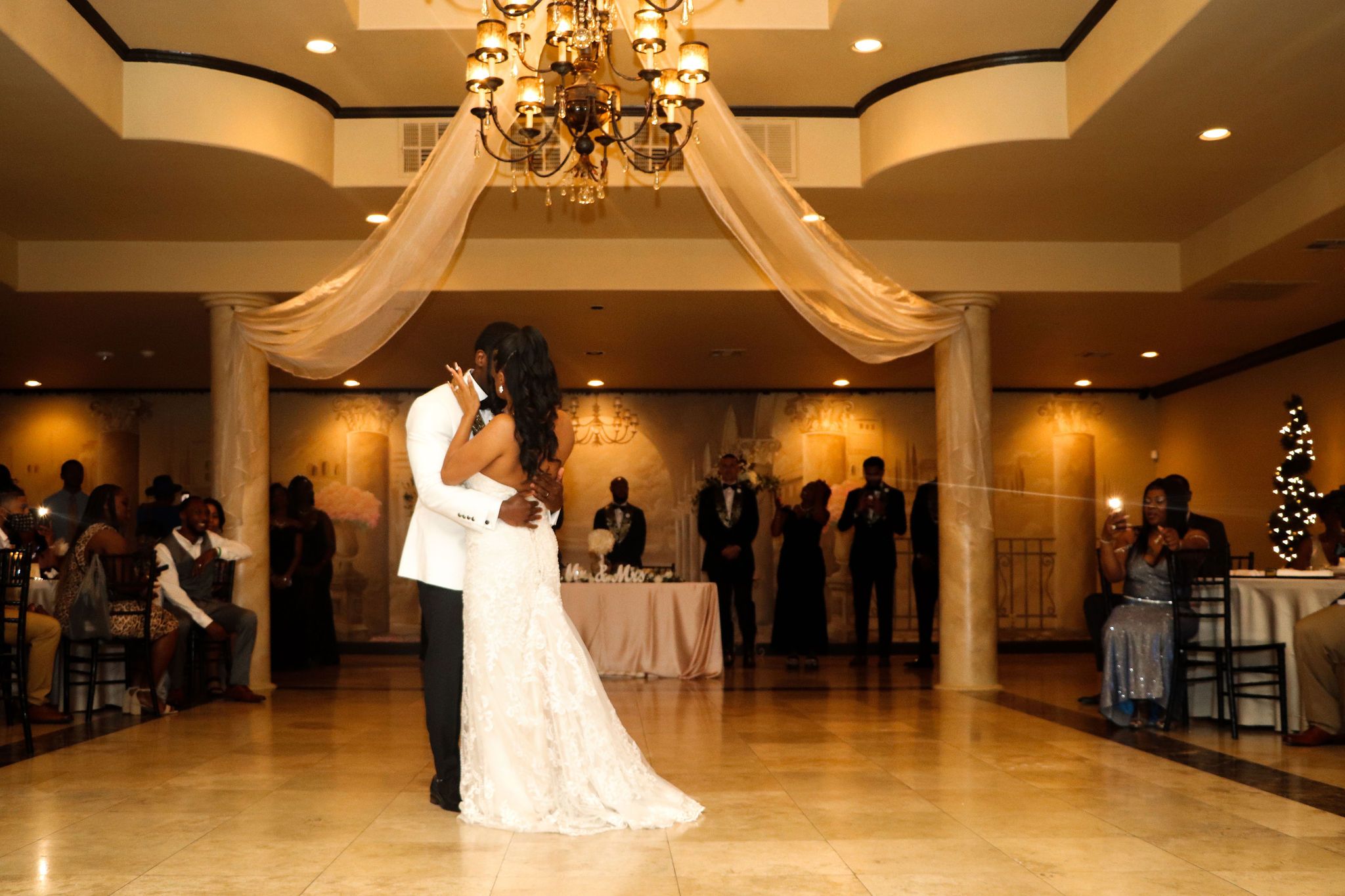 First dance of newlyweds under elegant chandelier and draped fabric in wedding reception ballroom.