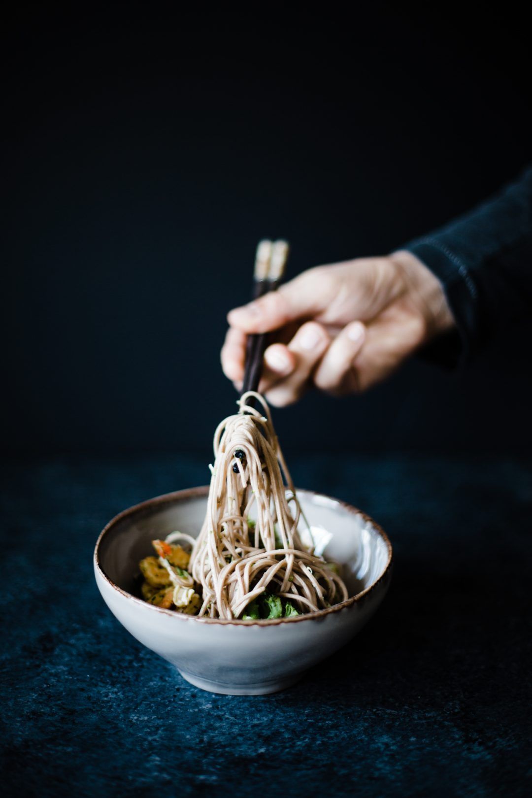 Hand lifting soba noodles with chopsticks from ceramic bowl against dark background.