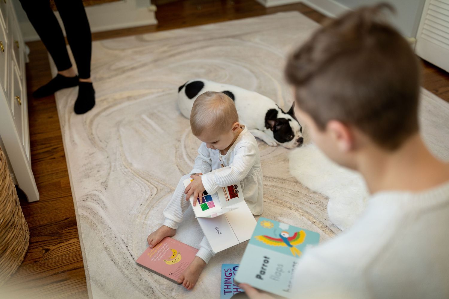A baby in white clothing explores books on the floor while being watched by someone.