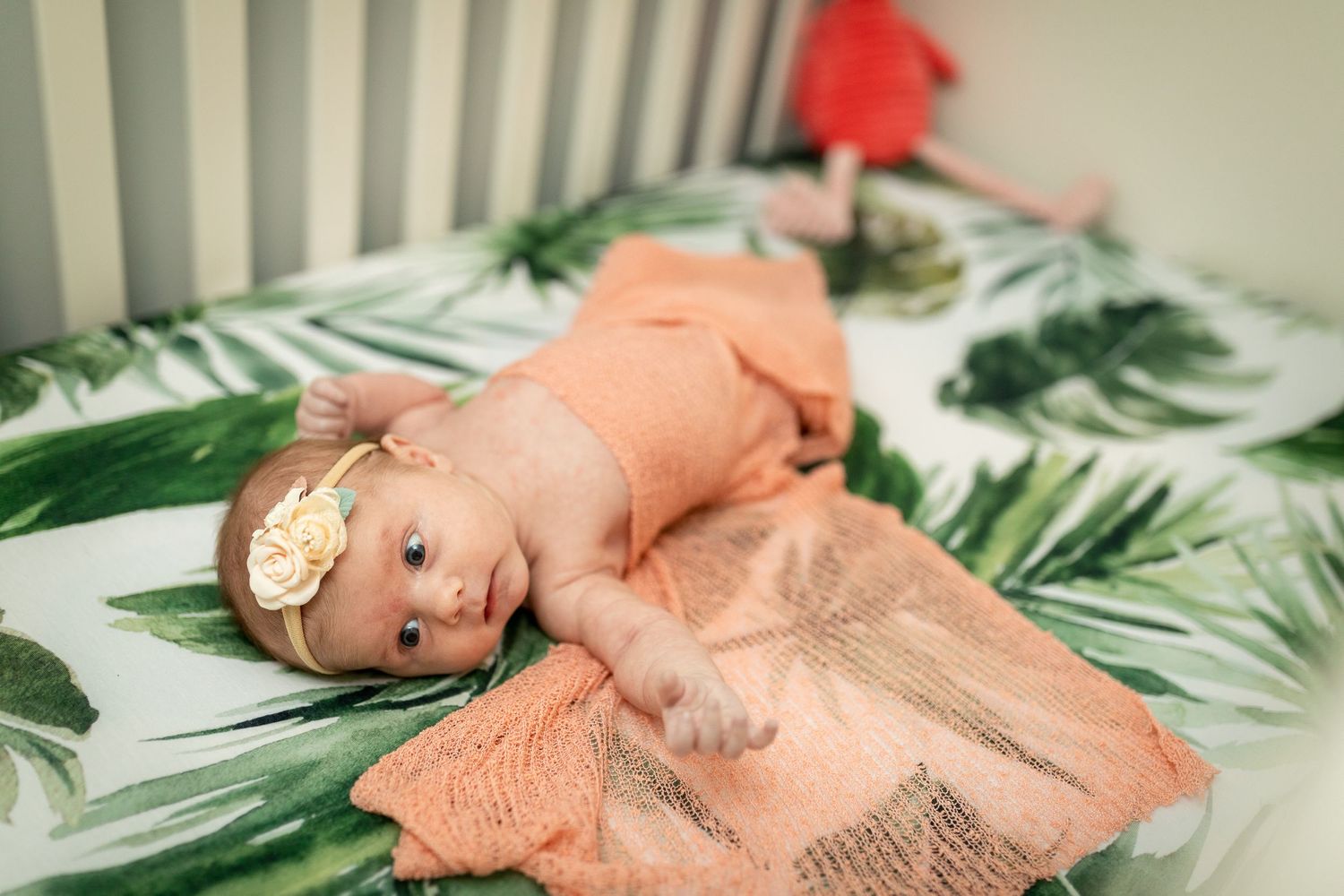 Baby in coral outfit lying on tropical leaf print backdrop.