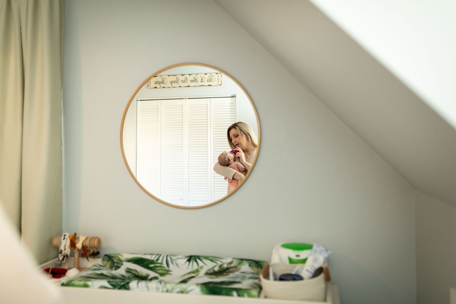 Modern nursery with sloped ceiling, white furniture, and oval mirror on gray walls.