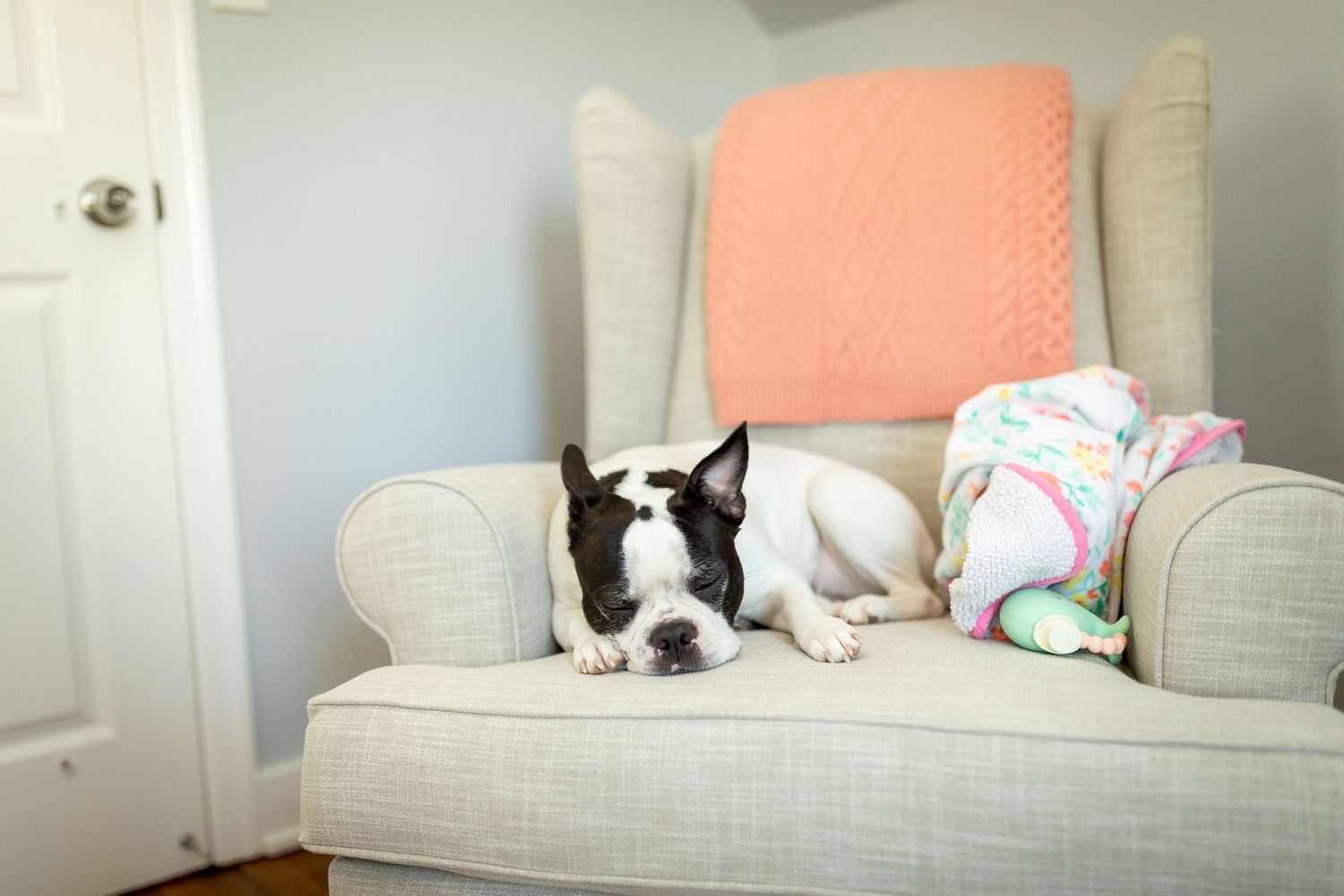 Nursery corner with white armchair, coral pillow, and cat watching from doorway.