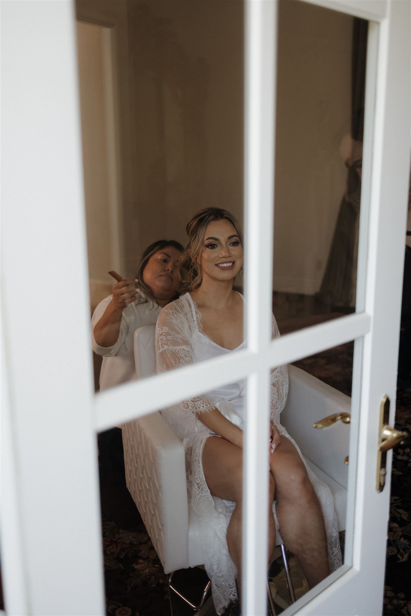 A wedding preparation scene viewed through a white-framed window pane showing intimate getting ready moments indoors.