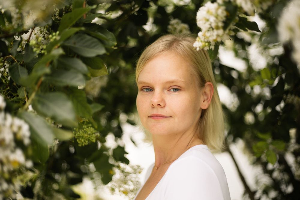 Portrait against white flowers and green leaves showing someone in a white top with blonde hair.