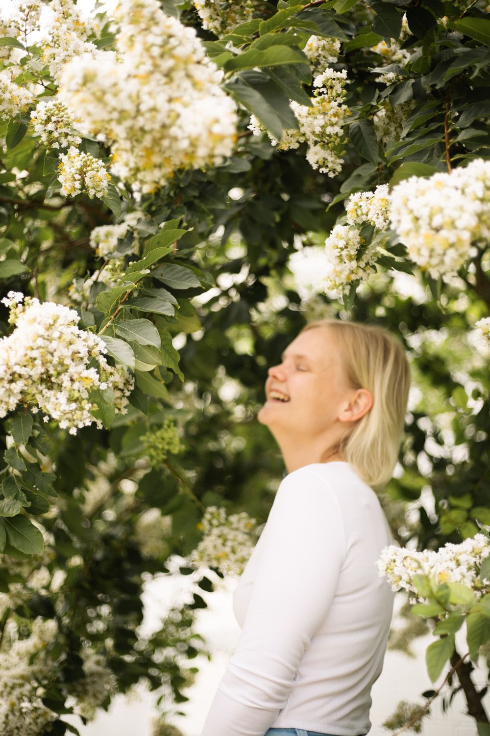 Person in white sweater laughing joyfully among blooming white flowers in a garden setting.