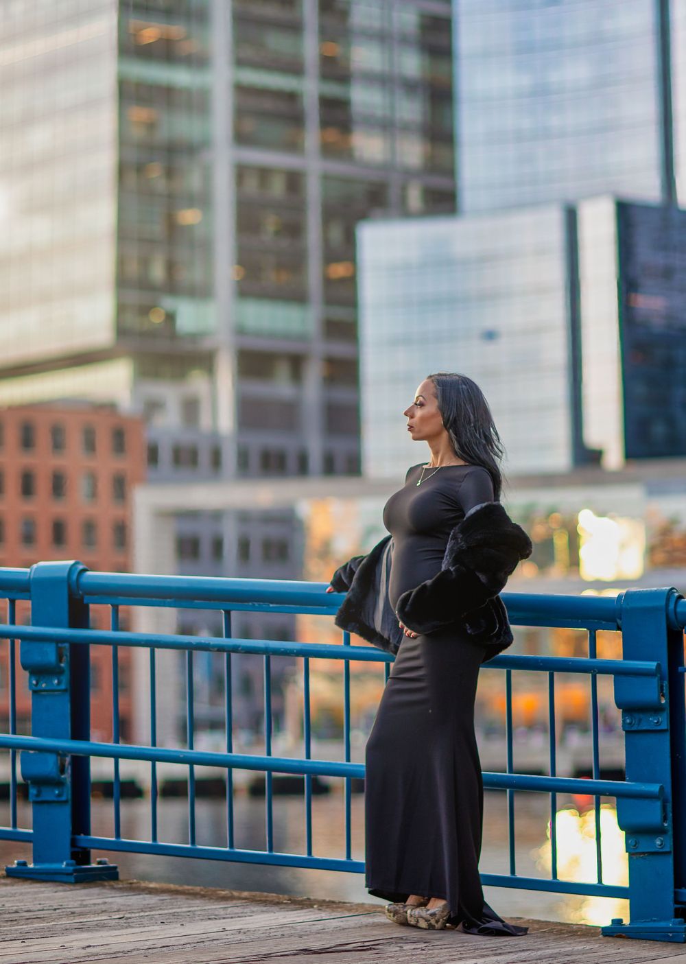 Silhouette of person in long dress standing on waterfront deck at sunset with city skyline.