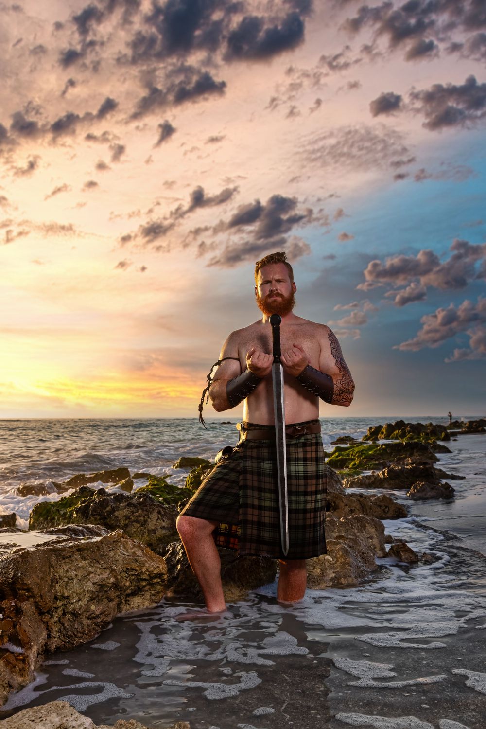 Man in traditional kilt poses dramatically on rocky shore at sunset.
