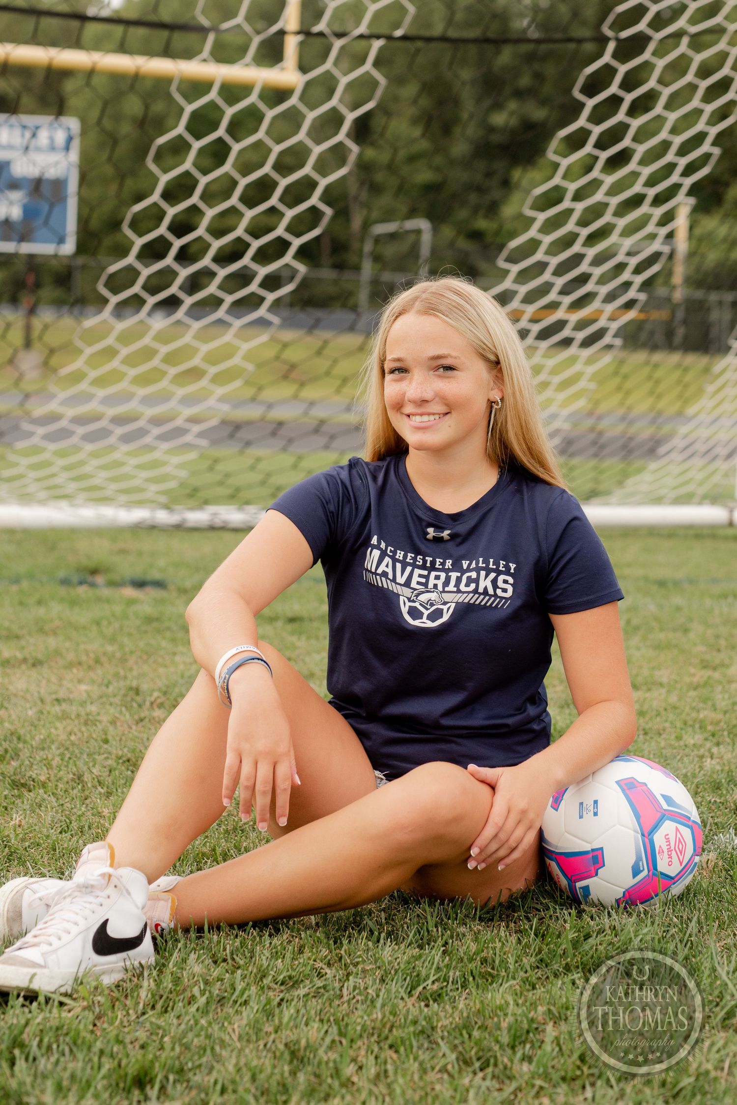 Soccer player poses with ball by goal net during outdoor sports photo session on grass field.
