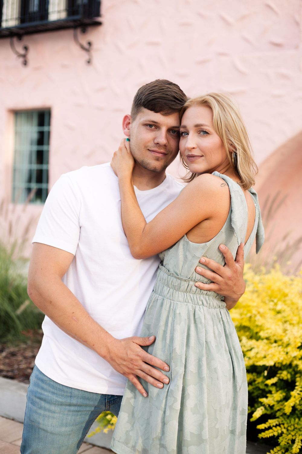 A couple poses intimately against a pink wall with decorative yellow bushes.