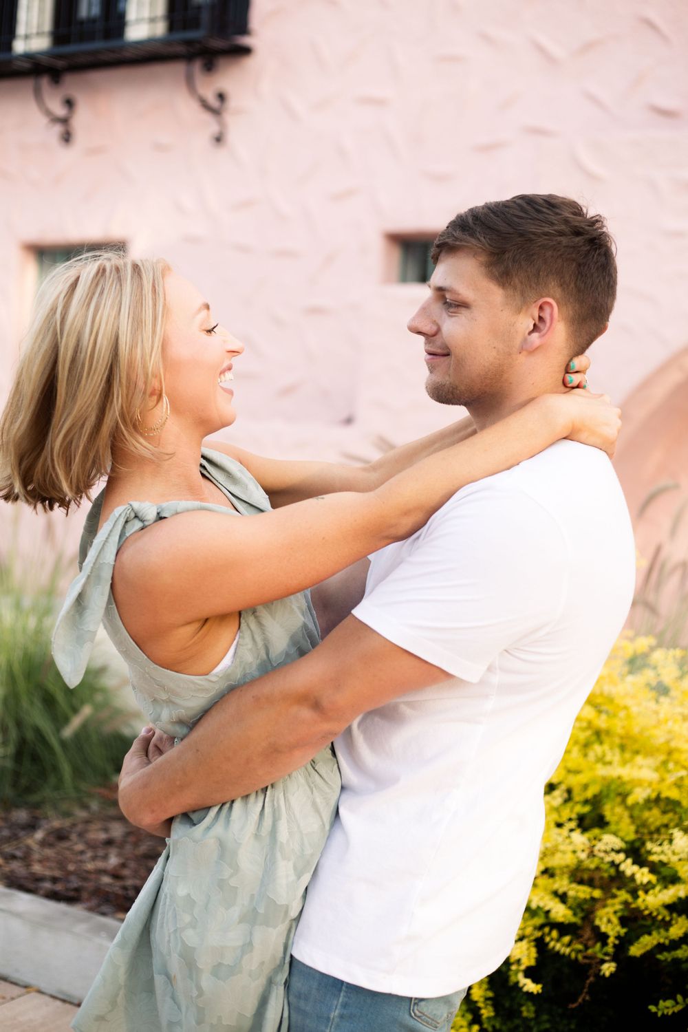 Romantic couple embracing and laughing together against a pink stucco wall.