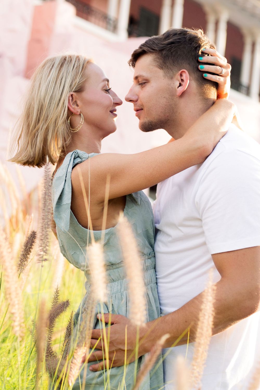 Romantic couple embracing among tall grasses with pink building backdrop.