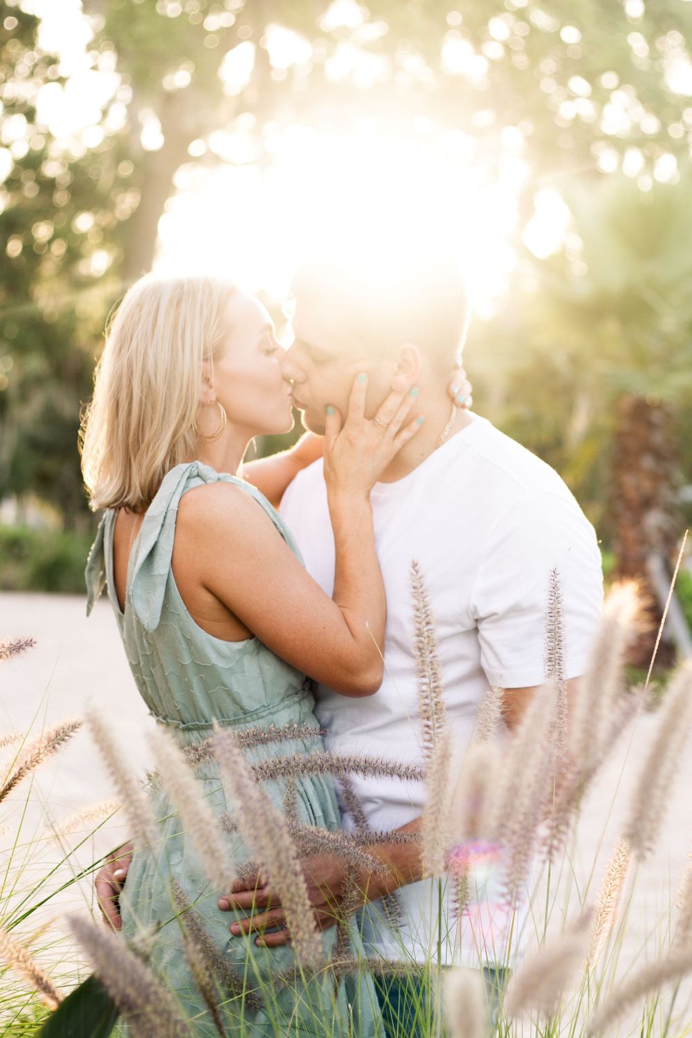 Couple sharing a sunset kiss framed by golden bokeh and wispy grasses.