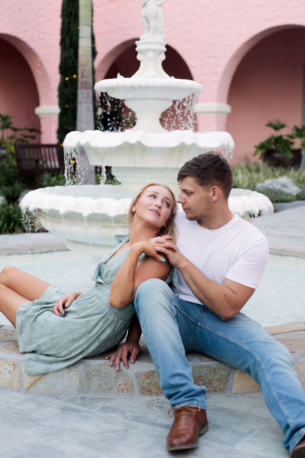 Couple relaxing by ornate white fountain against pink Mediterranean-style architecture.