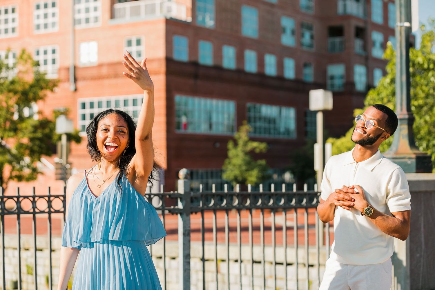 Joyful woman in blue dress raises her arm while man in white laughs beside her outdoors.