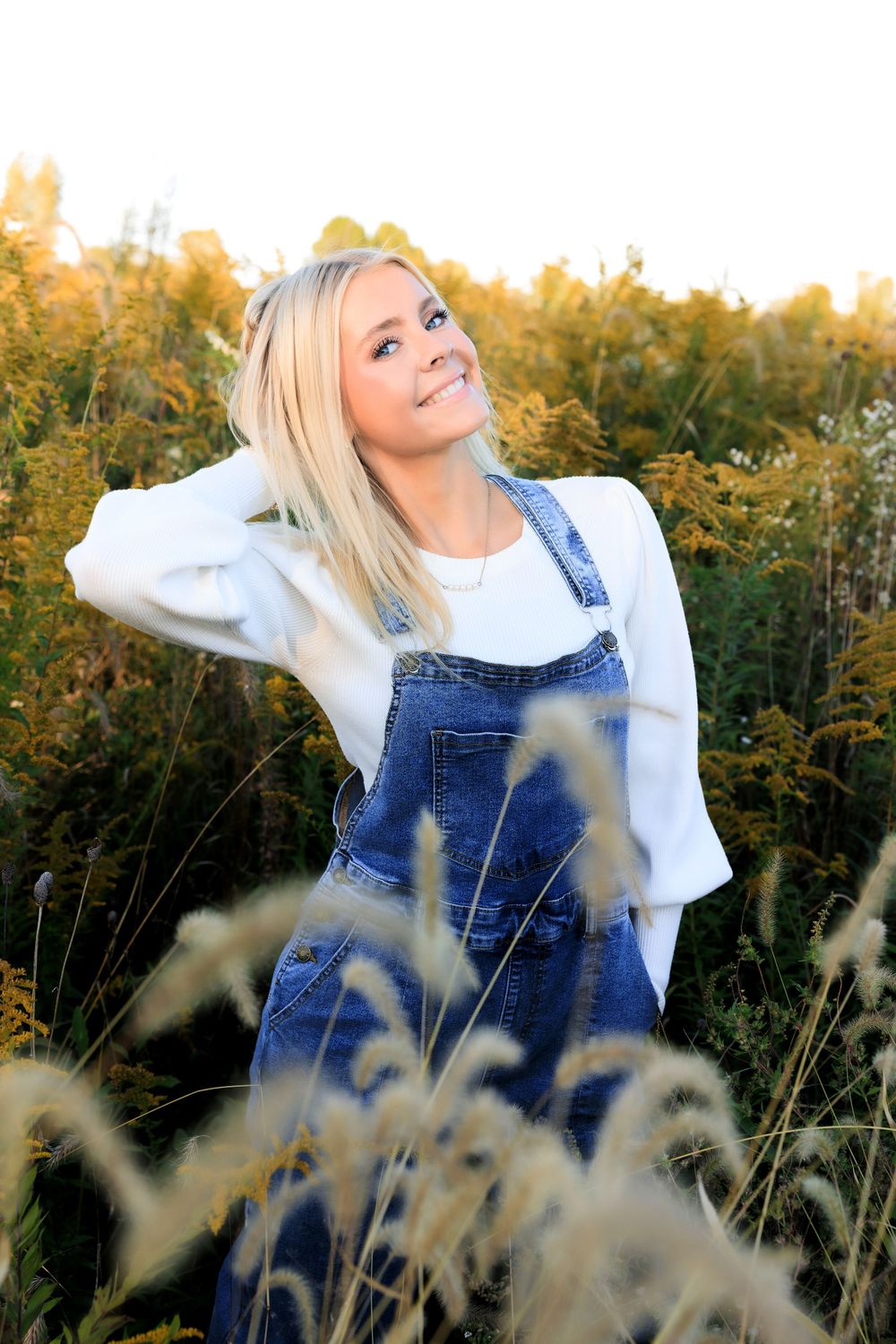 Playful pose in denim overalls and white sweater among tall autumn grasses.