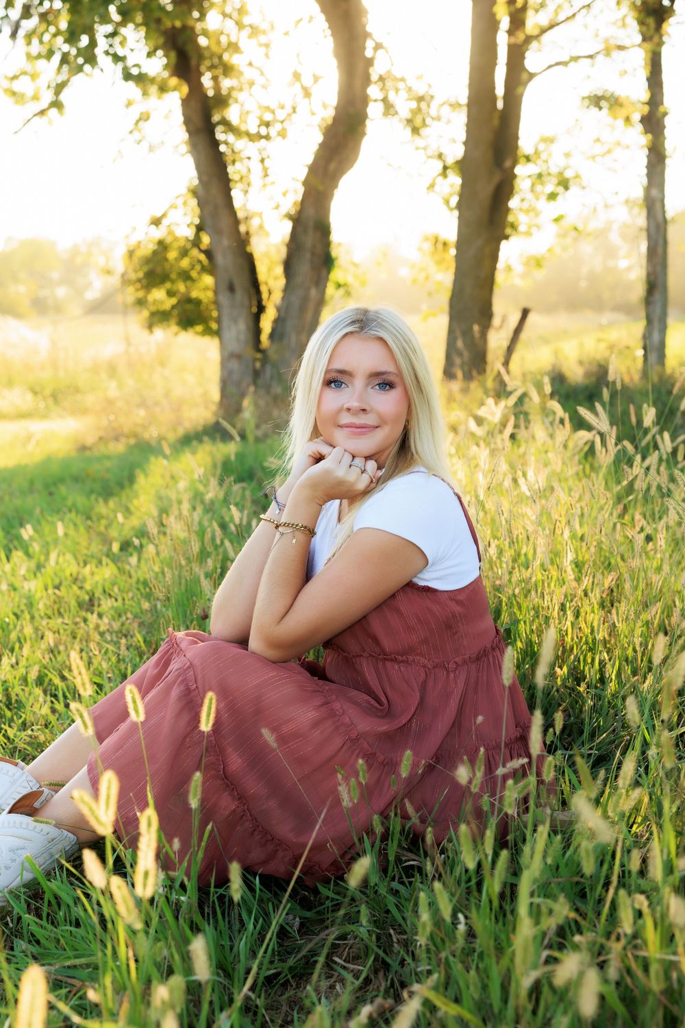 Peaceful outdoor portrait in burgundy dress sitting in sunlit grass near trees.
